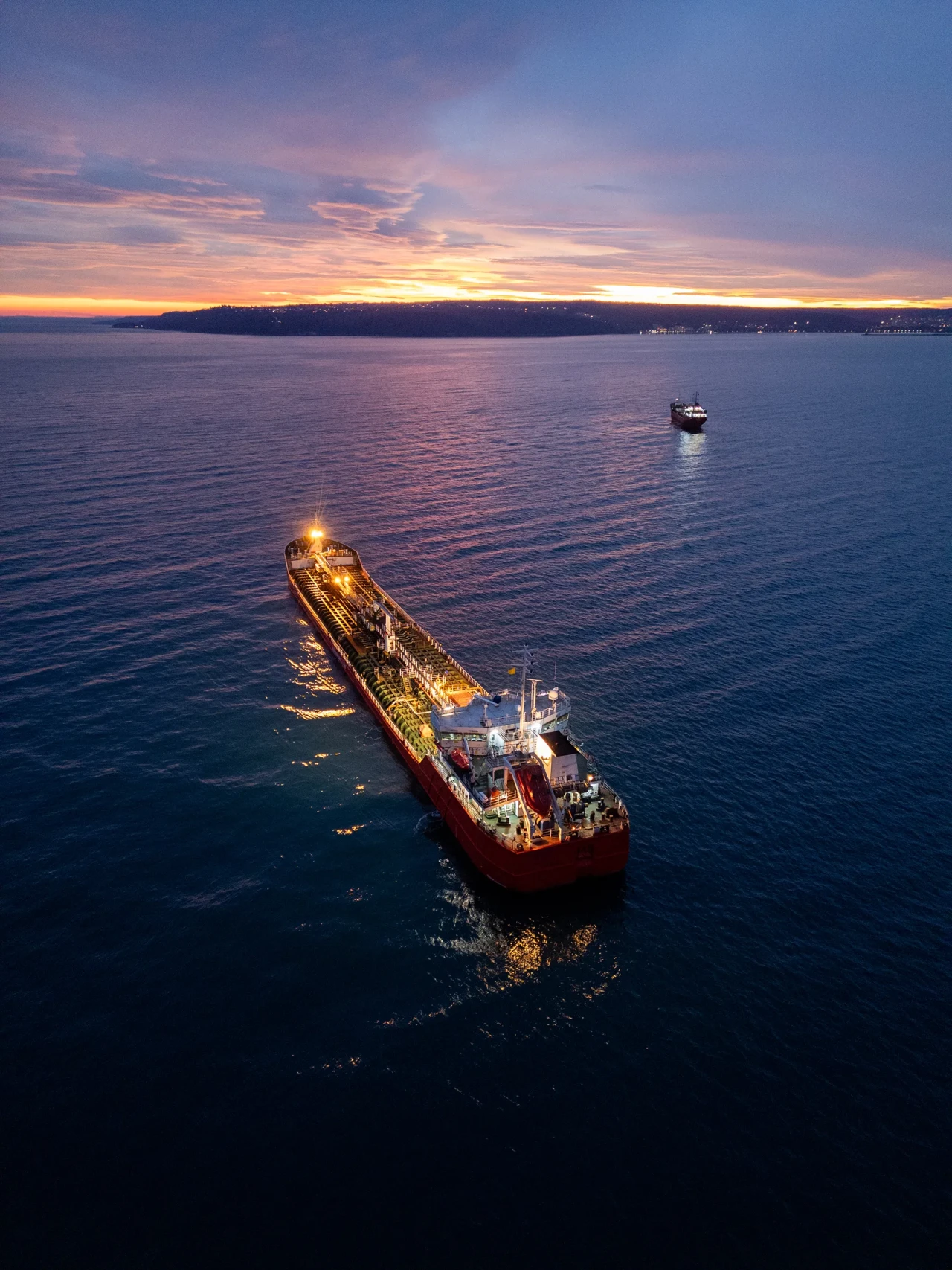 Aerial view of a brightly lit tanker ship on the sea during dusk, surrounded by open water and a glowing horizon.