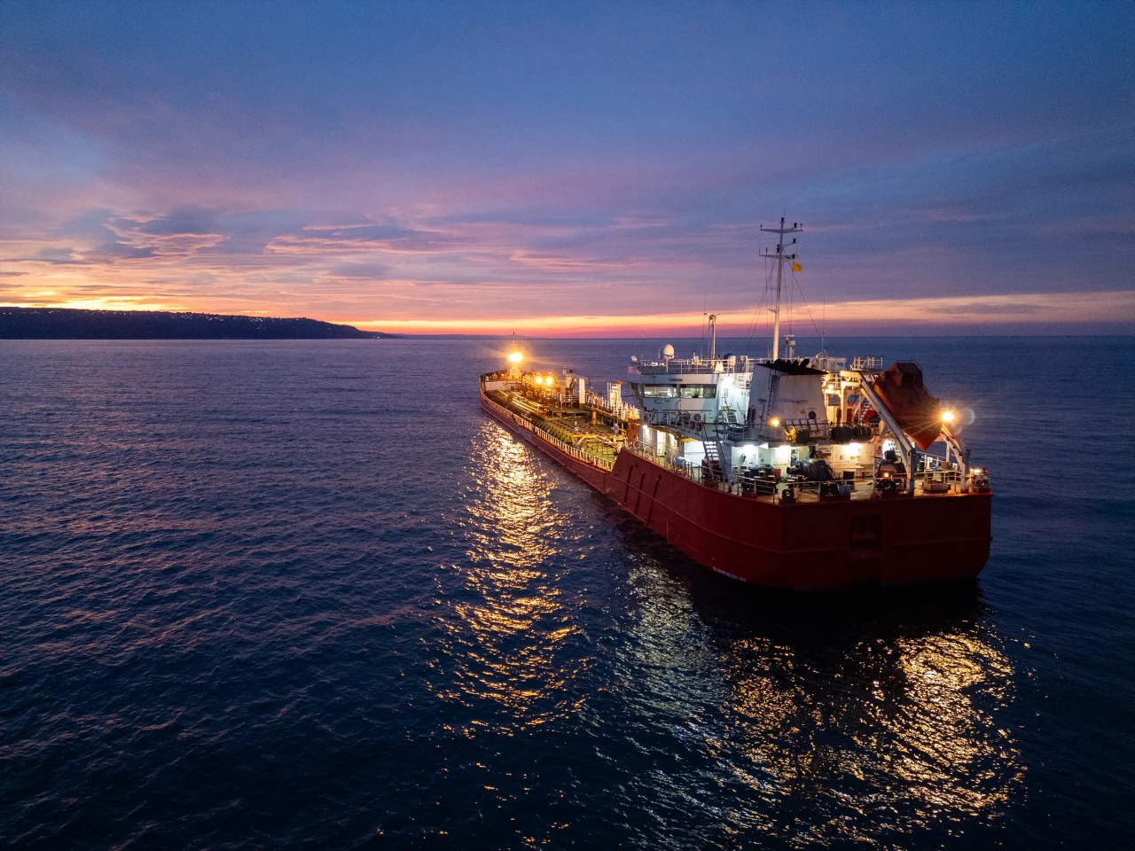 Aerial view of a brightly lit tanker ship on the sea during dusk, surrounded by open water and a glowing horizon.