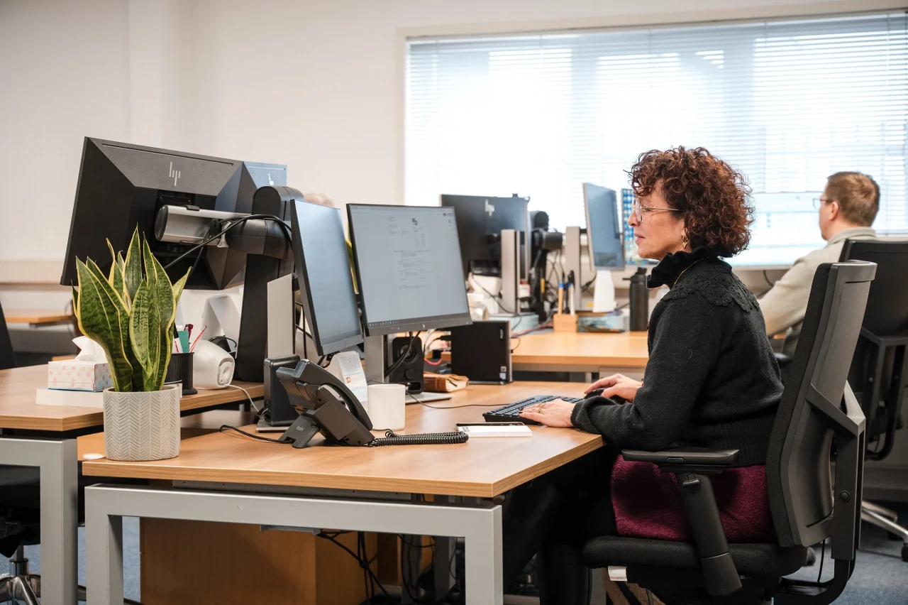 An Imatech employee works at her desk in a modern office, focusing on her screen while colleagues work at nearby workstations.
